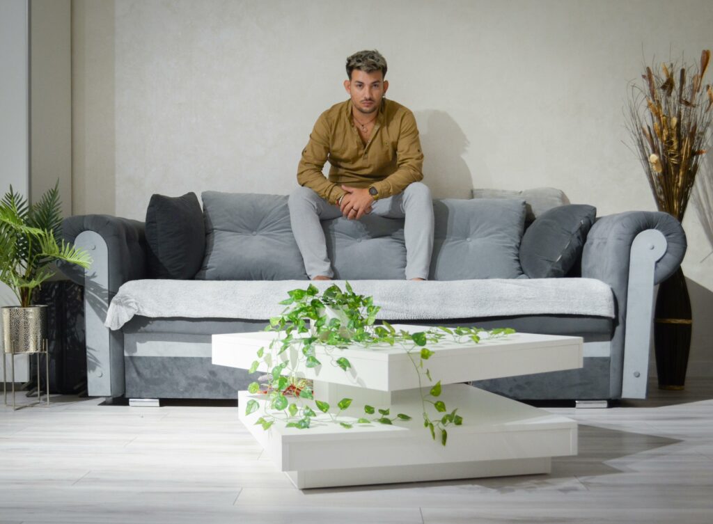 Man in brown shirt sitting on a grey sofa in a stylish indoor setting with plants and modern decor.